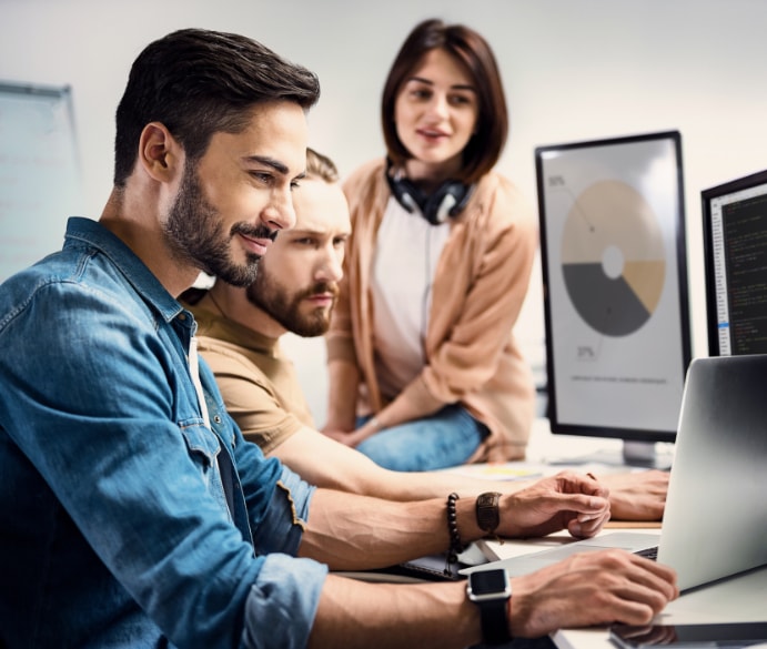 Three colleagues working together at a laptop beside monitors showing charts and code